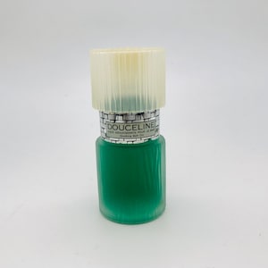 Op de afbeelding: Een groene glazen fles Douceline Soothing Bath Oil met een geribbelde, crèmekleurige dop. De fles heeft een getextureerd oppervlak en een zilveren etiket met zwarte tekst. De fles is gevuld met groene vloeistof.