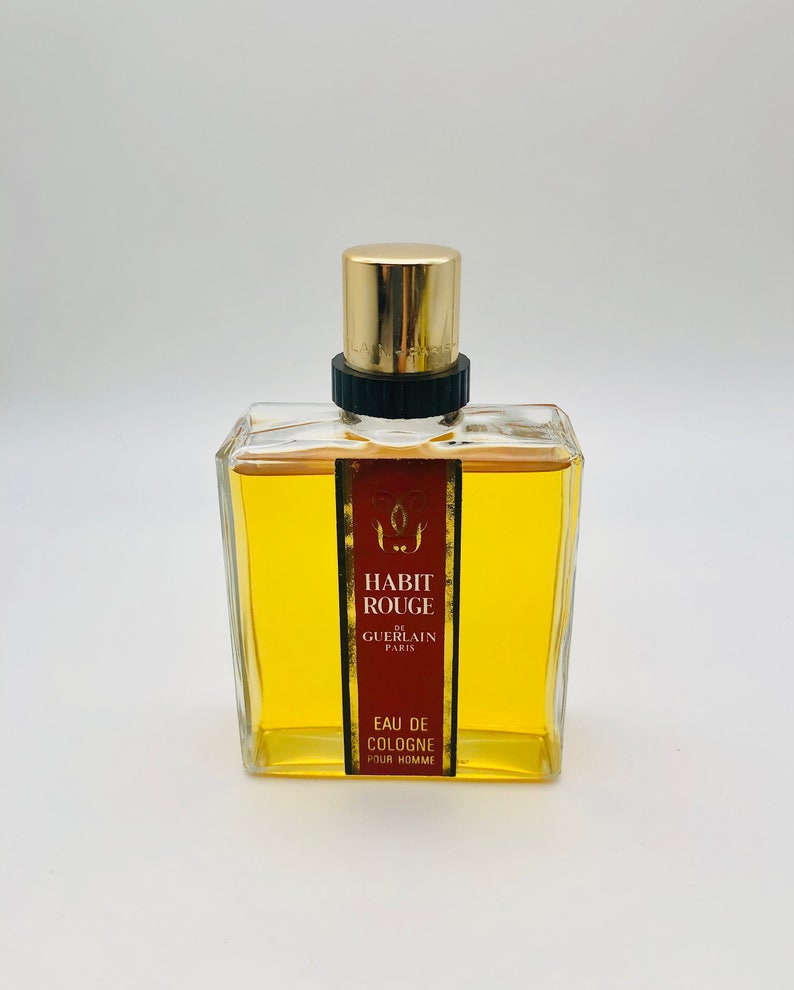 May include: A clear glass bottle of cologne with a gold cap and label. The label reads "Habit Rouge de Guerlain Paris Eau de Cologne Pour Homme".