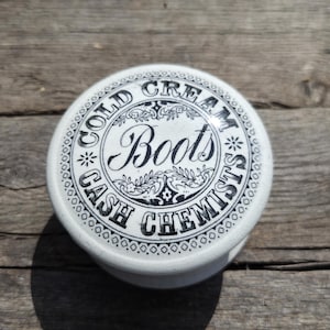 May include: A round white ceramic lid featuring black printed text that reads COLD CREAM BOOTS CASH CHEMISTS. The design includes decorative floral flourishes and a patterned border around the edge of the circular lid, creating a vintage aesthetic.
