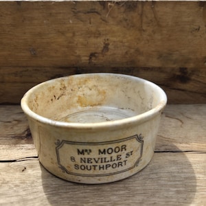 May include: A round, off-white ceramic pot with a weathered, stained finish. The exterior features a black rectangular border containing the printed text: MRS MOOR, 8 NEVILLE ST, SOUTHPORT. The vessel has a low profile and a wide, open top.