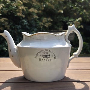 May include: A white ceramic teapot with a gold-trimmed rim and a handle. The side features a black graphic print that reads "Bobcross Brass Band 1905 Bazaar" with an illustration of a trumpet in the centre. The teapot has a curved spout.