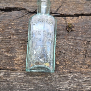 May include: A rectangular, light blue-green glass bottle with embossed text reading "PERALIA FOR THE COMPLEXION" and "PE & CO. SOUTH SHIELDS". The bottle is displayed on a weathered wooden surface, showcasing its vintage design.