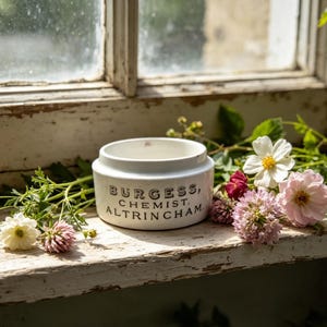 May include: A white ceramic ointment pot with a stepped rim and black printed text that reads BURGESS, CHEMIST, ALTRINCHAM. The cylindrical container is surrounded by a variety of small, colourful wildflowers including yellow, white, and pink blossoms.
