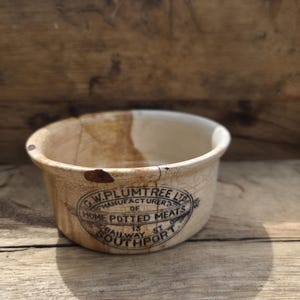 May include: A vintage off-white ceramic pot with a crackled glaze finish. The front features a black printed oval label reading G.W. PLUMTREE LTD MANUFACTURER OF HOME POTTED MEATS 13 RAILWAY ST SOUTHPORT. The vessel shows signs of age and wear.