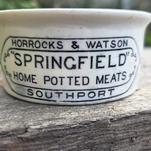May include: A white ceramic pot with black printed text that reads HORROCKS & WATSON, SPRINGFIELD, HOME POTTED MEATS, and SOUTHPORT. The container features a rounded rim and a decorative border surrounding the central text on the front side.
