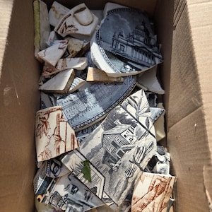 May include: A collection of broken ceramic shards featuring various transferware patterns in black, white, and brown. The pieces display detailed illustrations of architectural structures, landscapes, and botanical motifs, all contained within a cardboard box.