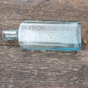May include: A rectangular, light blue glass bottle with the embossed text "HARGREAVES TIC MIXTURE CLITHEROE". The bottle is lying horizontally on a weathered, dark brown wooden surface.