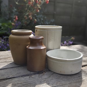 May include: A collection of four ceramic vessels including a tan rounded jar, a dark brown cylindrical bottle, a tall off-white fluted container, and a low white circular bowl. These items are arranged together, displaying varied shapes and earthy tones.
