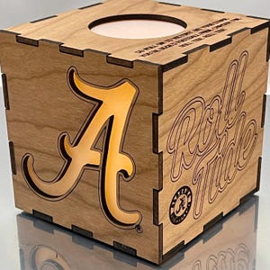 May include: A wooden box with a light brown finish. The box has a cutout of the letter "A" with a yellow glow behind it. The words "Roll Tide" are engraved on the side of the box. The box is a decorative item for fans of the University of Alabama.