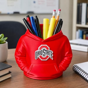 May include: A bright red desk organizer shaped like a miniature hoodie sweatshirt. The front features the Ohio State logo in white, red, and grey. The interior holds various pens, pencils, and highlighters in black, blue, and yellow colors.