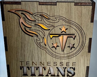 May include: A wooden box with a cutout design of the Tennessee Titans logo. The logo is illuminated from behind, creating a warm glow. The box is made of light brown wood and has a natural wood finish.