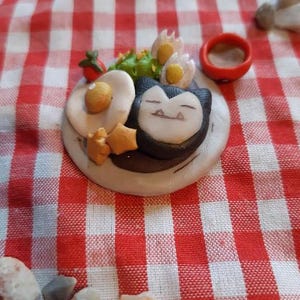 May include: A miniature clay figurine of a Pokemon character, Eevee, sitting on a plate with a fried egg, a small tomato, and a sprig of green. The plate is on a red and white checked tablecloth.