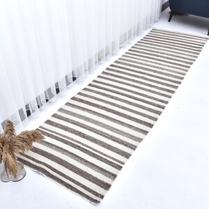Pode incluir: Um tapete corredor de listras brancas e marrons com um visual sutilmente desgastado. O tapete é feito de fibras naturais e tem um toque macio e texturizado.