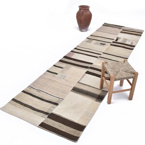 May include: A long, rectangular, off-white rug with brown stripes and patches. The rug is made of woven fabric and has a distressed look. A small wooden stool with a woven seat is in the foreground.