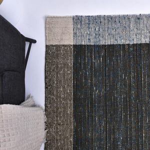 May include: A woven rug with a geometric design. The rug features a dark blue and black central section, a beige border, and a light blue top section. The rug is placed next to a dark gray armchair and a beige blanket.