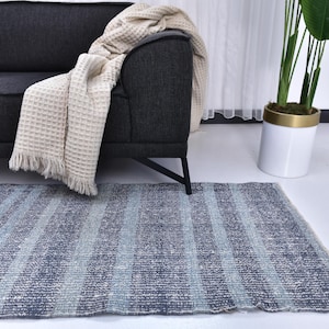 Op de afbeelding: Een rechthoekig tapijt met verticale strepen in blauw- en grijstinten. Het tapijt ligt op een witte vloer voor een donkergrijze bank met een crèmekleurige deken. Rechts staat een witte pot met een plant.