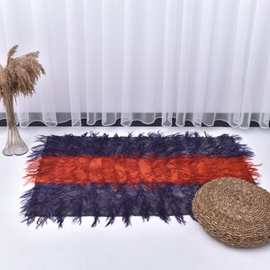 Peut inclure: Un tapis rectangulaire à texture touffue, avec une bande orange centrale entre deux sections bleu foncé. Un pouf rond et tressé marron est à proximité. Un grand vase avec des plantes séchées est à gauche.
