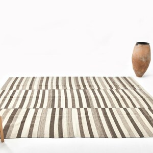 Op de afbeelding: Een rechthoekig tapijt met verticale strepen in bruin, beige en wit. Een kleine houten kruk met een geweven zitting staat ervoor. Er staat ook een terracotta vaas van ongeveer 60 cm hoog.