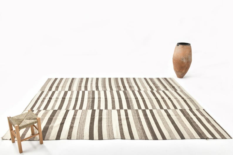 Op de afbeelding: Een rechthoekig tapijt met verticale strepen in bruin, beige en wit. Een kleine houten kruk met een geweven zitting staat ervoor. Er staat ook een terracotta vaas van ongeveer 60 cm hoog.