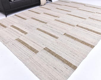 Può includere: Un grande tappeto rettangolare con sfondo color crema e strisce orizzontali marroni a spaziatura uniforme. Il tappeto è realizzato in materiale tessuto ed è posto su una superficie bianca. Sullo sfondo è visibile un divano grigio scuro.