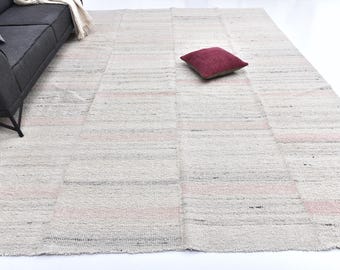 May include: A rectangular, off-white rug with horizontal stripes and pink accents. A dark gray sofa with a pillow and a burgundy pillow are also visible. The rug is made of woven material.