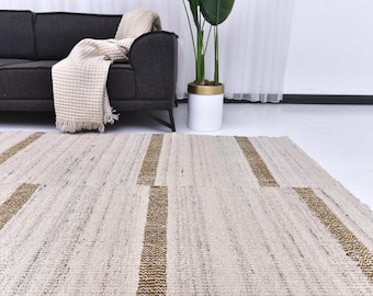 Può includere: Un tappeto neutro con una trama intrecciata e un design geometrico. Il tappeto presenta strisce alternate di beige e un colore marrone dorato più scuro. Il tappeto è posto di fronte a un divano grigio scuro.
