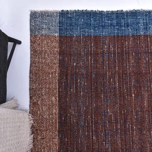Op de afbeelding: Een rechthoekig tapijt met een geweven textuur. Het tapijt heeft een colorblock-ontwerp met een blauw bovenstuk, een bruin middenstuk en een bruine verticale streep. Het tapijt is gemaakt van geweven materiaal.