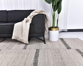 May include: A modern living room scene featuring a dark gray sofa with a cream-colored throw blanket. A large, patterned rug in neutral tones with black accents covers the floor. A white planter with a gold rim holds a tall green plant.
