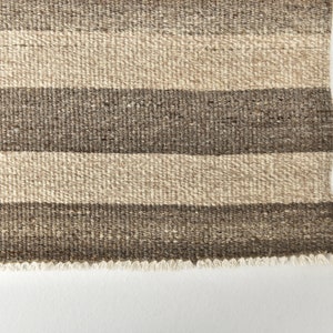 May include: A close-up of a woven rug with brown and beige stripes. The rug has a fringed edge.