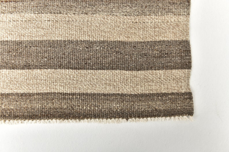 May include: A close-up of a woven rug with brown and beige stripes. The rug has a fringed edge.