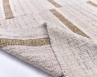 Può includere: Primo piano di un tappeto beige e marrone tessuto con un motivo a righe. Il tappeto ha una superficie strutturata e un angolo piegato, che rivela il lato inferiore. Le strisce marroni sono uniformemente distanziate.