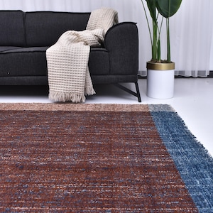 Op de afbeelding: Een rechthoekig vloerkleed met een geweven textuur. Het vloerkleed heeft een colorblock-ontwerp met een bruin midden, een beige rand en een blauwe rand. Een donkergrijze bank en een plant in een pot staan op de achtergrond.