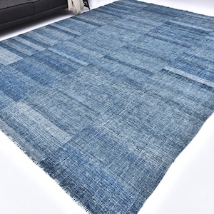 Op de afbeelding: Een groot, rechthoekig blauw tapijt met een patchwork-ontwerp. Het tapijt heeft verschillende blauwtinten, waardoor een textuur ontstaat. Het tapijt ligt op een witte ondergrond, met op de achtergrond een donkergrijze bank.