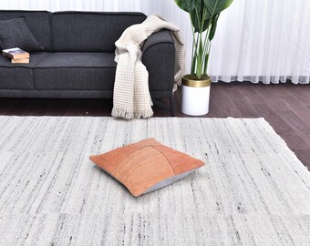 May include: An orange and tan square pillow with a geometric design rests on a white and gray striped rug. A dark gray sofa with pillows and a cream-colored throw blanket is in the background. A potted plant is also visible.