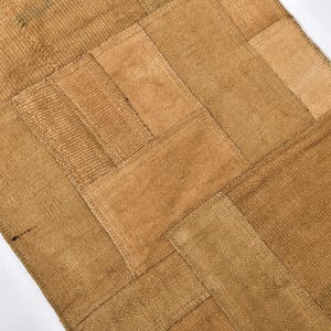 Puede incluir: Una pieza textil rectangular compuesta por múltiples parches de tela de color tostado y marrón claro cosidos en un patrón de cuadrícula. La tela muestra una textura tejida visible con diversos tonos de marrón tierra en toda la superficie.