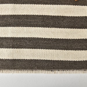 May include: A brown and white striped rug with a fringe border. The rug is made of a woven material and has a subtle texture.