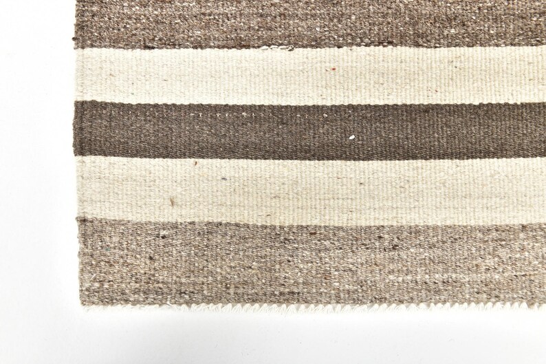 Op de afbeelding: Close-up van een geweven tapijt met een gestreept patroon. Het tapijt heeft afwisselende banden van lichtbeige, donkerbruin en middenbruin. De textuur van het tapijt is zichtbaar, met een licht gerafelde rand.