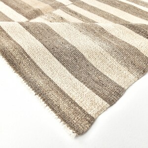May include: Close-up of a woven rug with alternating stripes of beige and brown. The rug has a fringed edge.
