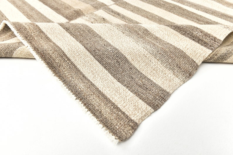 May include: Close-up of a woven rug with alternating stripes of beige and brown. The rug has a fringed edge.