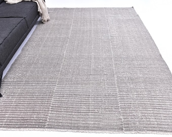 May include: A large, rectangular area rug in shades of gray and white. The rug has a textured, striped pattern and is placed on a white surface. A dark gray sofa is partially visible in the upper left corner.