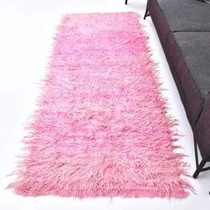 Könnte beinhalten: Ein langer, flauschiger rosa Teppich mit einer zotteligen Textur. Der Teppich liegt auf einer weißen Oberfläche.