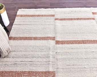 May include: A rectangular, woven rug with a neutral color palette. The rug features horizontal bands of a warm, reddish-brown color against a background of off-white and gray. The texture appears slightly coarse, suggesting a natural fiber.