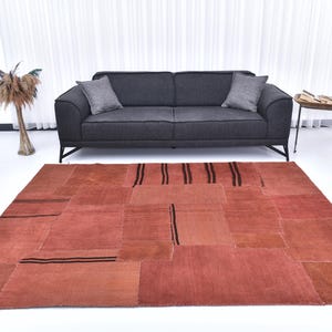 May include: A patchwork rug with a red-brown color and black stripes. The rug is made of different pieces of fabric sewn together. It is placed in front of a gray sofa in a modern living room.