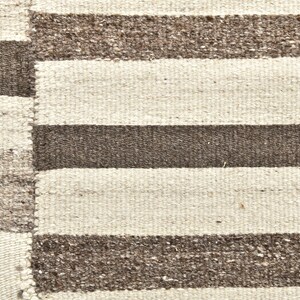 Op de afbeelding: Close-up van een geweven tapijt met een gestreept patroon. Het tapijt heeft afwisselende banden van lichtbeige en donkerbruin. De textuur van het tapijt is zichtbaar, met een enigszins ruwe, handgemaakte uitstraling. Het tapijt is waarschijnlijk gemaakt van natuurlijke vezels.