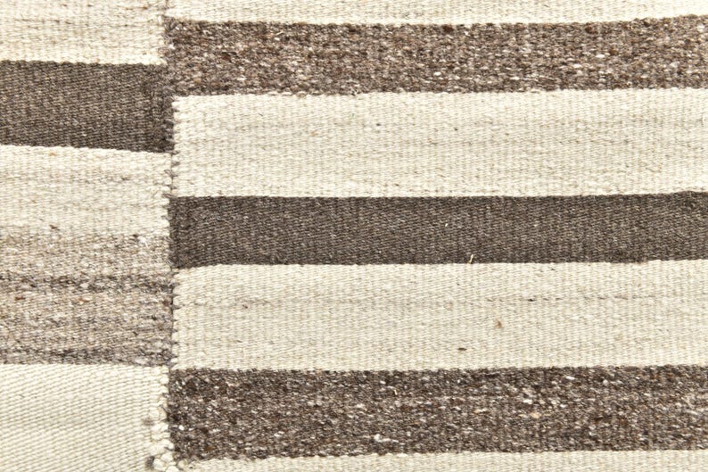 Op de afbeelding: Close-up van een geweven tapijt met een gestreept patroon. Het tapijt heeft afwisselende banden van lichtbeige en donkerbruin. De textuur van het tapijt is zichtbaar, met een enigszins ruwe, handgemaakte uitstraling. Het tapijt is waarschijnlijk gemaakt van natuurlijke vezels.