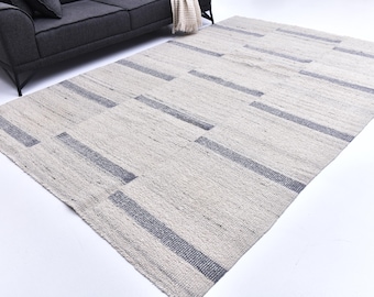 May include: A rectangular, off-white rug with a geometric pattern of gray stripes. The rug is made of a woven material and is placed on a white floor. A gray sofa is partially visible in the background.