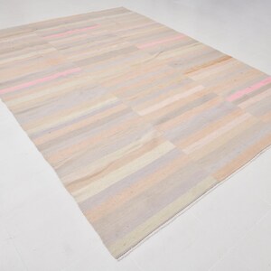 K&ouml;nnte beinhalten: Ein rechteckiger Teppich mit einem gestreiften Muster in Beige-, Rosa- und Graut&ouml;nen. Der Teppich hat einen wei&szlig;en Fransenrand.