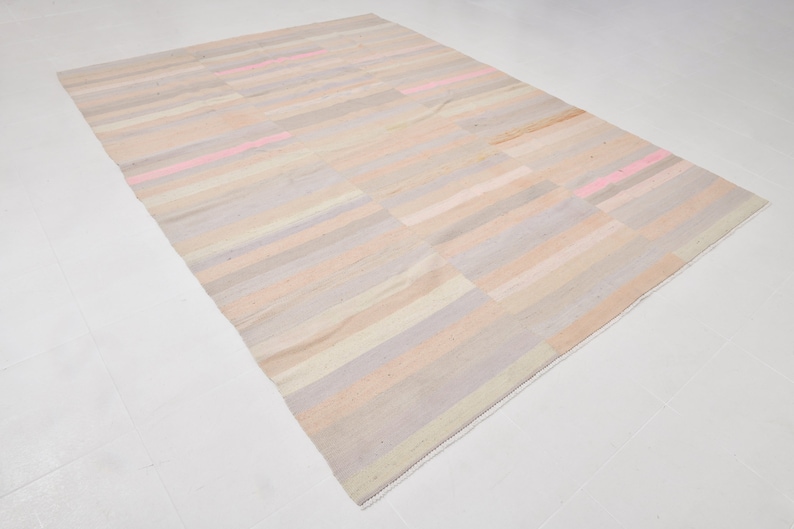 K&ouml;nnte beinhalten: Ein rechteckiger Teppich mit einem gestreiften Muster in Beige-, Rosa- und Graut&ouml;nen. Der Teppich hat einen wei&szlig;en Fransenrand.