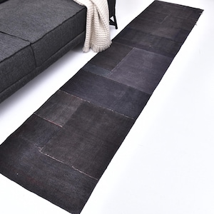 Pode incluir: Um tapete preto retangular e longo com um design de patchwork. O tapete é feito de várias tonalidades de tecido preto, criando uma aparência texturizada. É colocado em um chão branco ao lado de um sofá cinza escuro. O tapete tem aproximadamente 3 metros de comprimento.