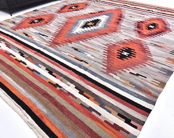 May include: A large, rectangular rug with a geometric pattern. The rug features a central design of diamond shapes in orange, gray, and black, surrounded by stripes of various colors including red, brown, and blue. The rug is made of woven fabric.